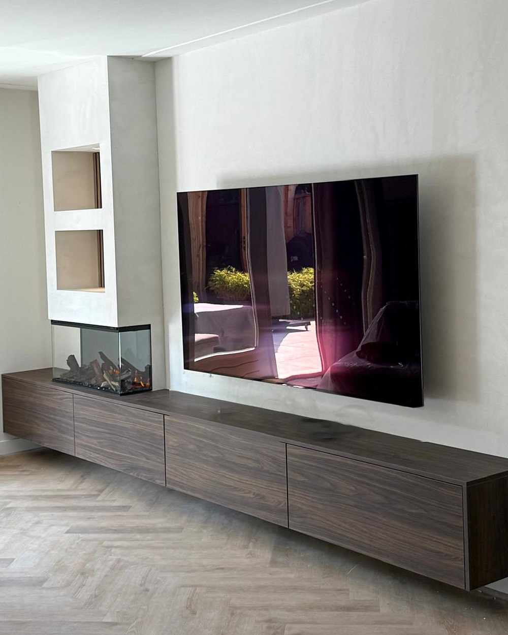 De wand vanaf de zijkant, met de brandende driezijdige haard, de televisie die de tuin weerspiegelt en het zwevende dressoir over de volle lengte