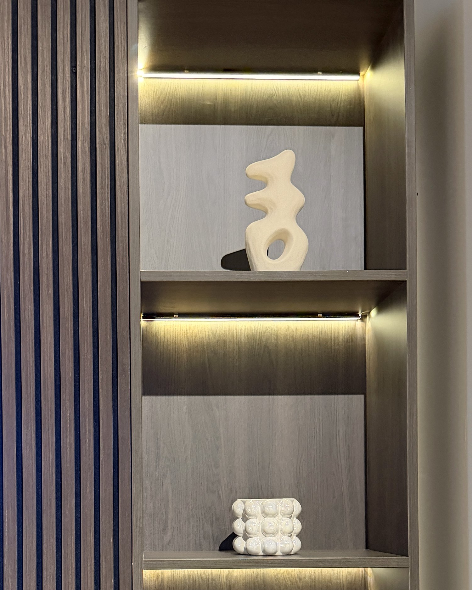Twee verlichte vakken in de kast van dichtbij, met een doorlopende lichtlijn boven elke plank en het ritme van de lamellen ernaast